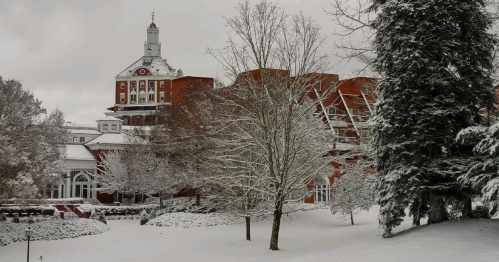 A snowy landscape featuring a historic red brick building with a clock tower, surrounded by bare trees and winter scenery.