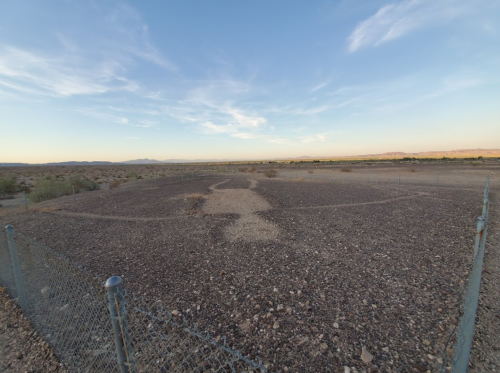 A fenced area in a desert landscape, featuring rocky ground and sparse vegetation under a clear blue sky.