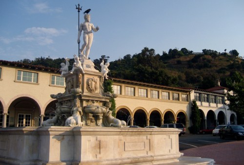 A white statue of a figure holding a staff atop a fountain, surrounded by sculptures, with buildings and hills in the background.