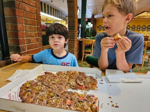 Two boys sit at a table with a half-eaten pizza box, one eating a slice and the other looking at the camera.