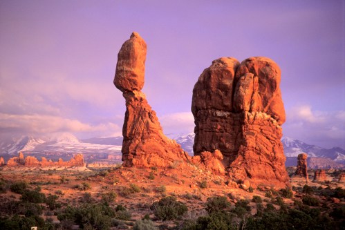 A striking rock formation with a tall, balanced stone against a colorful sky and distant mountains.