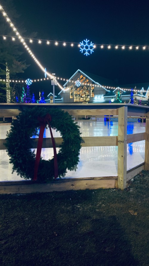A festive wreath hangs on a wooden railing, with an ice skating rink and colorful lights in the background at night.