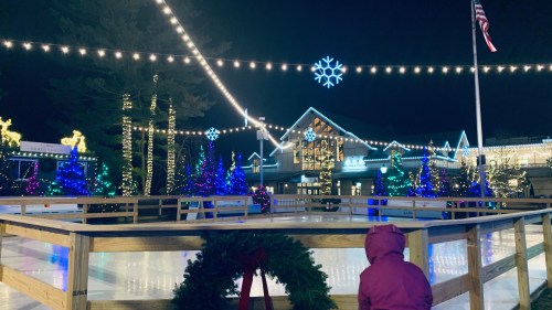 A child in a pink coat stands by an ice rink, surrounded by festive lights and decorated trees at night.