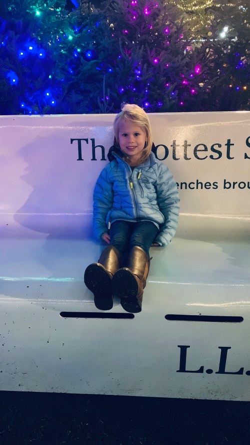A young girl with blonde hair sits on a bench, smiling, with colorful lights in the background.