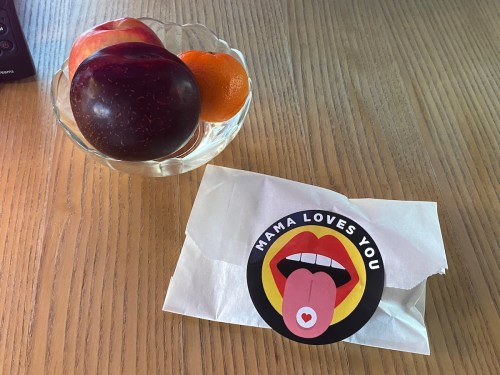 A bowl of fruit with an apple and a tangerine next to a wrapped item featuring a sticker that says "MAMA LOVES YOU."