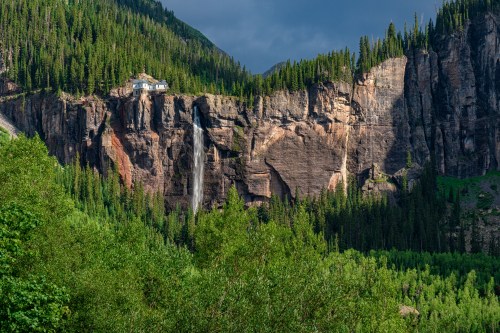 A waterfall cascades down a rocky cliff surrounded by lush green trees, with a house perched on the cliffside above.