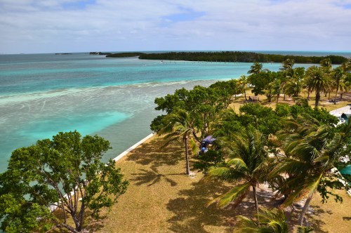 A scenic view of turquoise waters, lush greenery, and palm trees along a coastline under a partly cloudy sky.