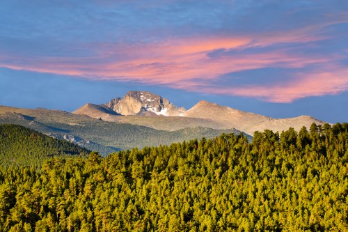 A majestic mountain peak rises above a dense forest, illuminated by a colorful sunset sky.