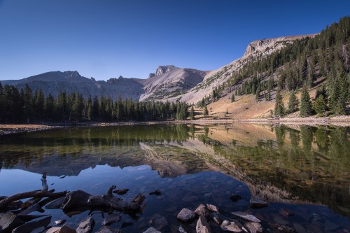 A serene mountain landscape reflecting in a calm lake, surrounded by trees and rocky terrain under a clear blue sky.