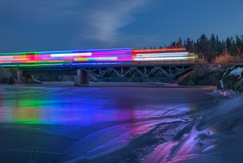 A colorful train moves across a bridge at dusk, reflecting vibrant lights on the icy river below.