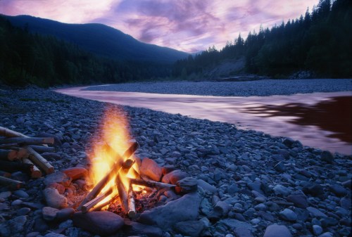 A campfire burns on a rocky riverbank, with mountains and a colorful sunset in the background.