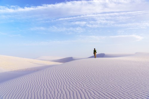 A person stands on rippled white sand dunes under a blue sky with wispy clouds.