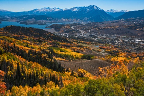 A panoramic view of colorful autumn foliage, mountains, and a lake, with a highway winding through the valley below.
