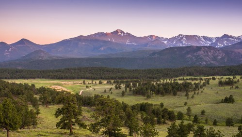 A scenic view of mountains at sunset, with lush green fields and trees in the foreground.