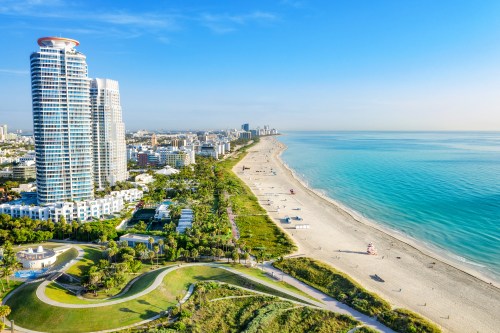 Aerial view of a sunny beach with tall buildings, green parks, and calm turquoise waters along the coastline.