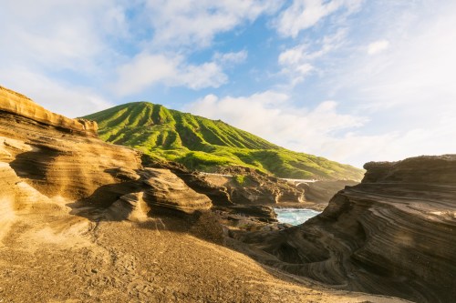 A scenic view of a green mountain rising above rocky coastal formations under a blue sky with scattered clouds.