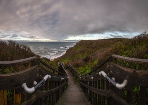 A wooden staircase leads down to a coastal view with waves crashing against the shore under a cloudy sky.