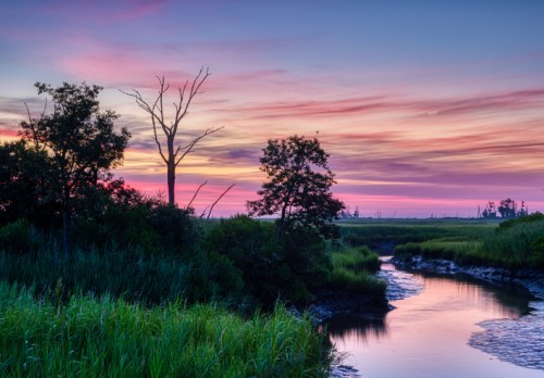 A serene landscape at sunset, featuring a winding stream, lush greenery, and colorful skies with pink and purple hues.