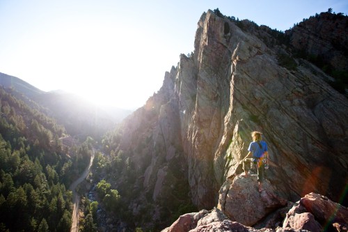 A climber stands on a rocky outcrop overlooking a valley and mountains, with the sun shining in the background.