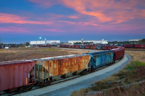 A curved train track with weathered freight cars under a colorful sunset sky. Industrial buildings are in the background.
