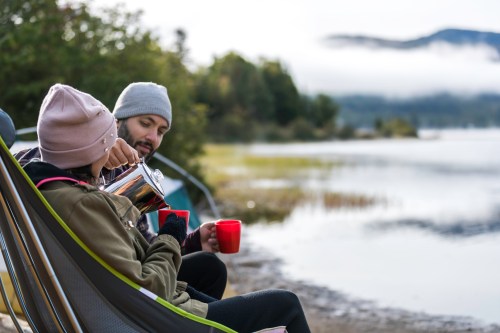 A couple sits by a lake, enjoying coffee from a pot, surrounded by nature and misty mountains.