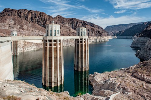A dam with two tall towers stands over a calm reservoir, surrounded by rocky mountains and a clear blue sky.