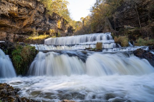 A serene waterfall cascades over rocky steps, surrounded by lush greenery and sunlight filtering through trees.