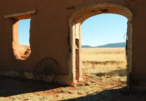 A weathered adobe wall with two openings, revealing a vast landscape and a wooden wagon wheel nearby.