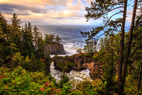 A scenic coastal view with rocky cliffs, lush trees, and waves crashing against the shore under a colorful sky.