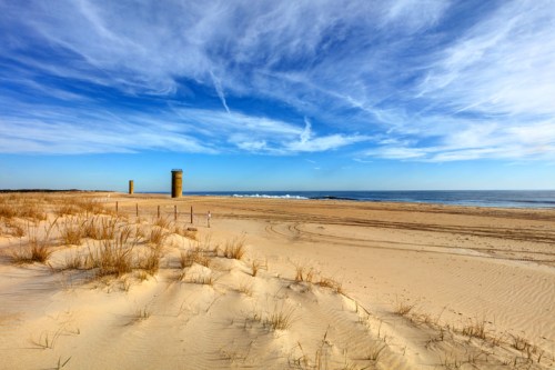 A sandy beach with tall structures in the distance, under a blue sky with wispy clouds and gentle ocean waves.