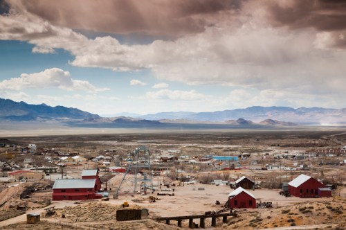 A panoramic view of a desert town with red buildings, mountains in the background, and dramatic clouds overhead.