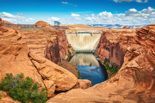 A scenic view of a dam surrounded by red rock formations and a river, under a blue sky with fluffy clouds.