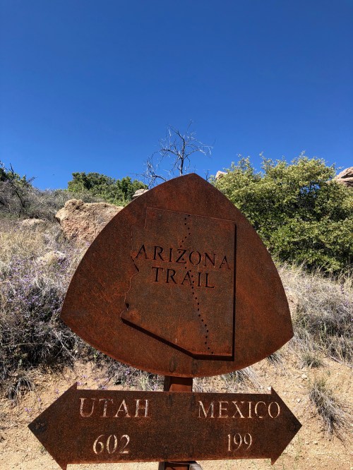 Rusty trail sign for the Arizona Trail, indicating distances to Utah (602 miles) and Mexico (199 miles) against a blue sky.