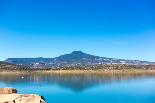 A serene lake reflects a mountain under a clear blue sky, surrounded by lush greenery and rocky terrain.
