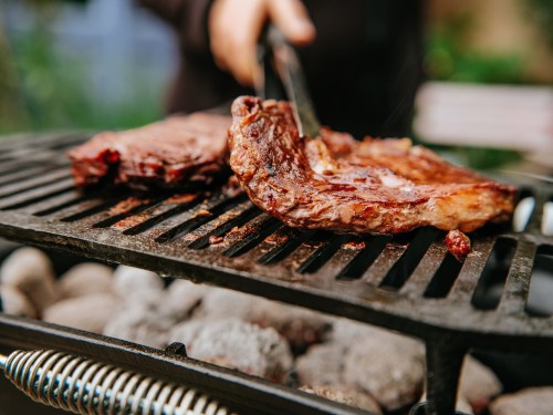 A close-up of grilled meat on a barbecue grill, with smoke rising and a hand using tongs to handle the meat.