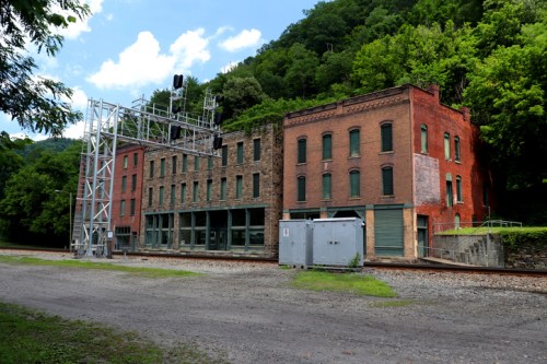 Abandoned brick buildings alongside a railway track, surrounded by lush green hills under a blue sky.