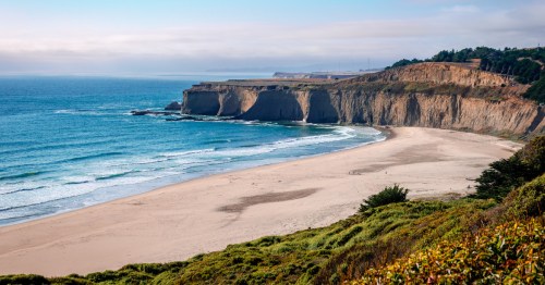 A scenic view of a sandy beach with cliffs and gentle waves under a clear blue sky.