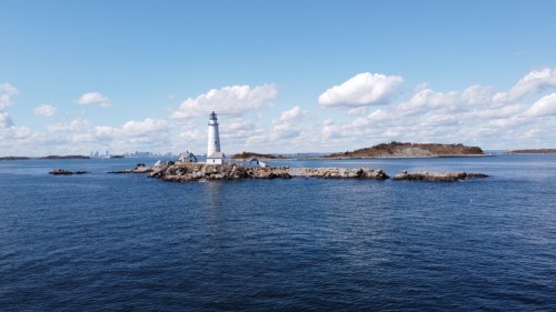 A lighthouse stands on a rocky island surrounded by calm blue waters, with a distant city skyline under a partly cloudy sky.
