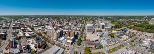 Aerial view of a cityscape featuring buildings, roads, and green spaces under a clear blue sky.