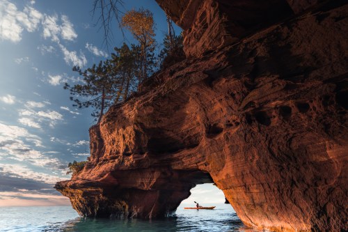 A kayaker paddles through a natural rock arch, surrounded by trees and calm waters under a colorful sky.