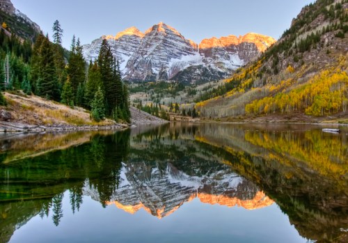 A serene lake reflects snow-capped mountains and autumn trees at sunrise, creating a picturesque landscape.