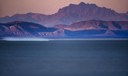 A distant view of mountains at dusk, with a lone vehicle on a flat, reflective surface in the foreground.