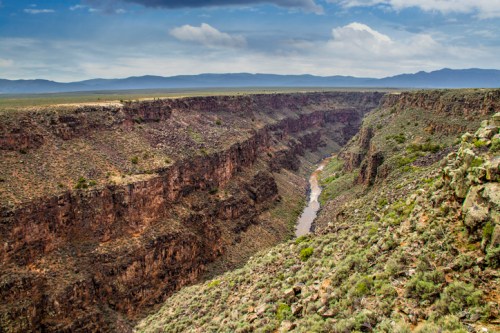 A vast canyon with steep rocky walls and a winding river below, surrounded by green vegetation and distant mountains.
