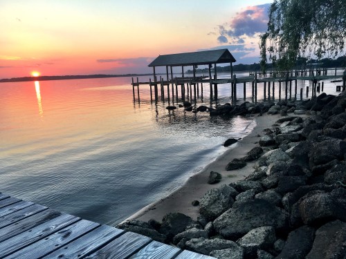 Sunset over calm water, with a wooden pier and rocky shoreline, creating a serene lakeside scene.