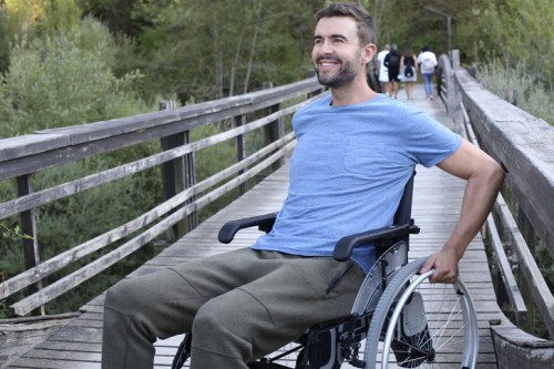 A smiling man in a wheelchair on a wooden bridge, surrounded by greenery and people walking in the background.