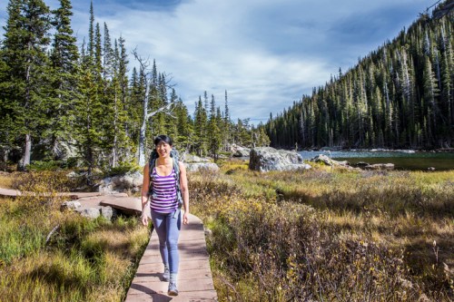 A woman walks along a wooden path through a scenic landscape of trees and mountains by a lake.