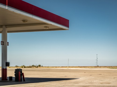 A deserted gas station under a clear blue sky, with an empty lot and distant power lines in the background.