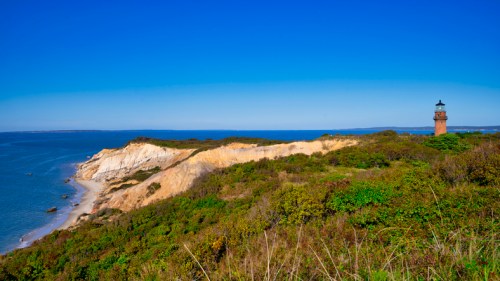 A scenic view of a lighthouse on a cliff overlooking the ocean, with lush greenery and clear blue skies.