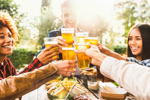 A group of friends toasting with glasses of beer outdoors, enjoying food and a sunny atmosphere.