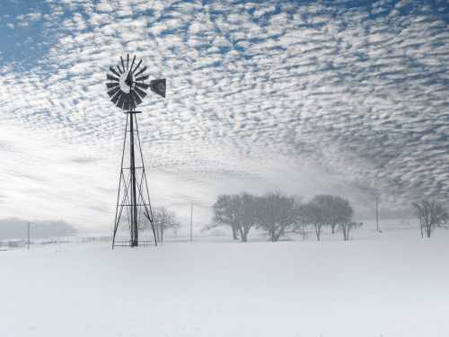 A windmill stands in a snowy landscape under a sky filled with textured clouds. Trees are faintly visible in the background.